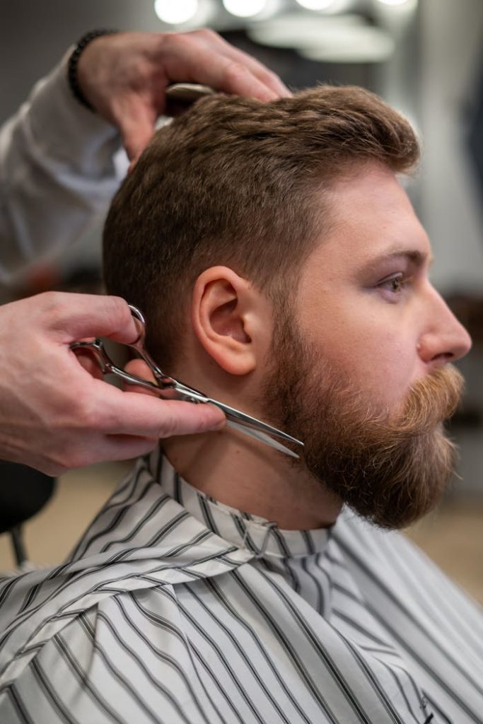 A side view of a barber trimming a bearded mans hair in a barber shop.